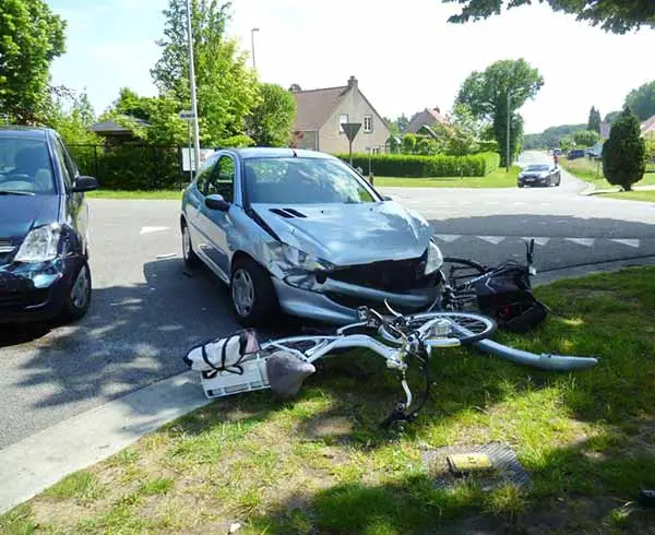 a silver car with a crushed front end and a wrecked bicycle lying on the grass beside it after a collision