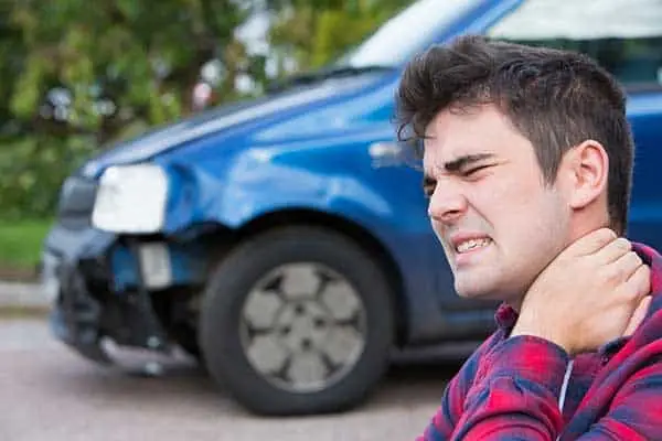 Man gripping his neck in pain in front of a blue car with a wrecked front end