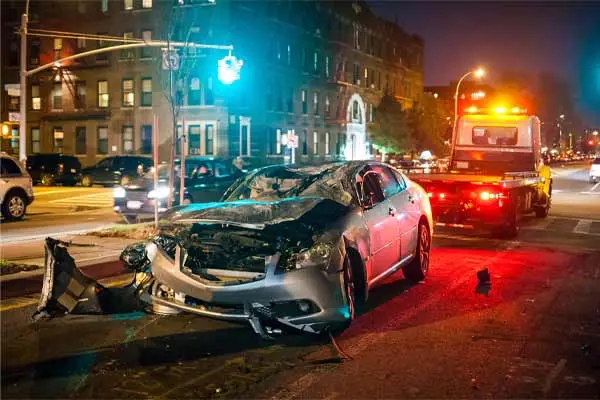 A wrecked car at a city intersection at night with a tow truck behind it