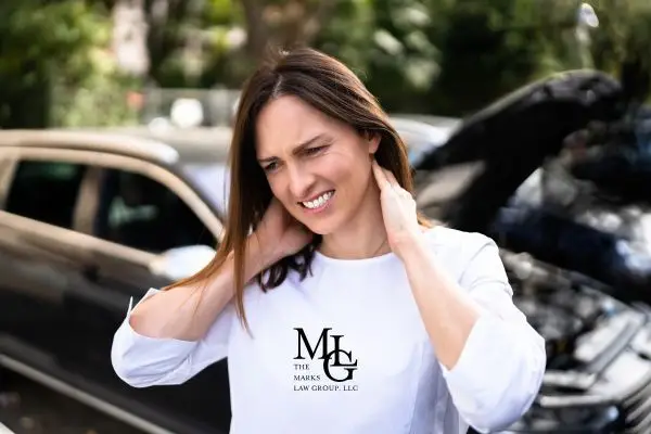 Woman in a white shirt holding her neck in pain in front of a damaged car with the hood up.