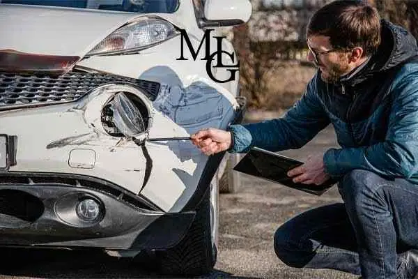 A man crouching to inspect a dented front bumper while holding a tablet