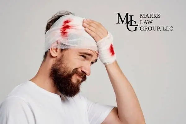 Bearded man wincing with a bloodstained bandage around his head and hand