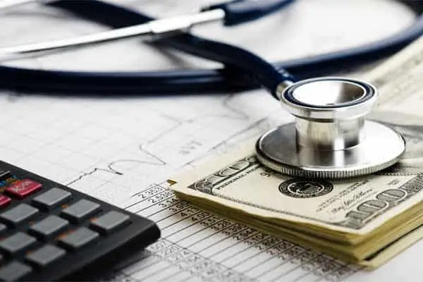 A stethoscope on a stack of hundred-dollar bills beside a calculator and medical charts