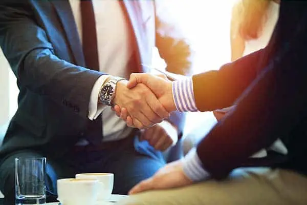 Two people in business dress shaking hands across a table with coffee cups