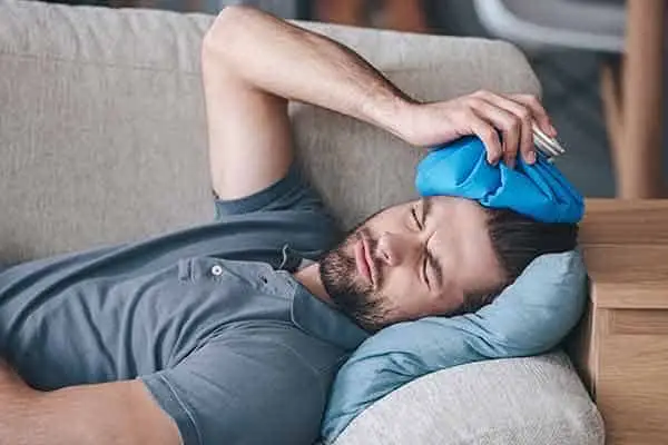 bearded man lying on a couch holding a blue ice pack to his head
