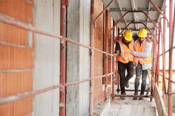 two workers in hard hats and orange vests walking along scaffolding at a building site