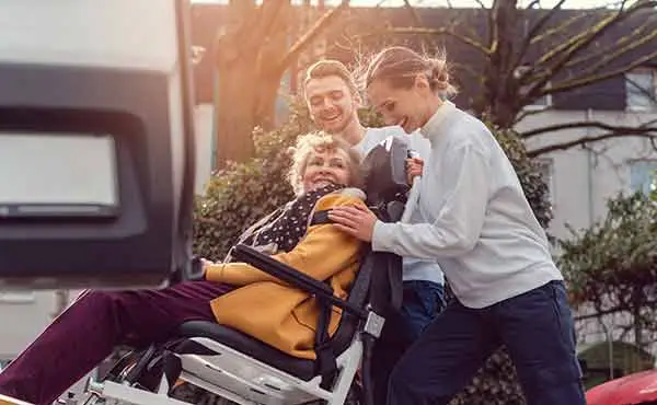two caregivers loading a smiling older woman in a wheelchair into a van
