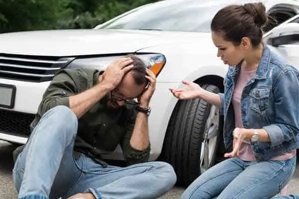 man sitting on the road holding his head beside a white car and woman