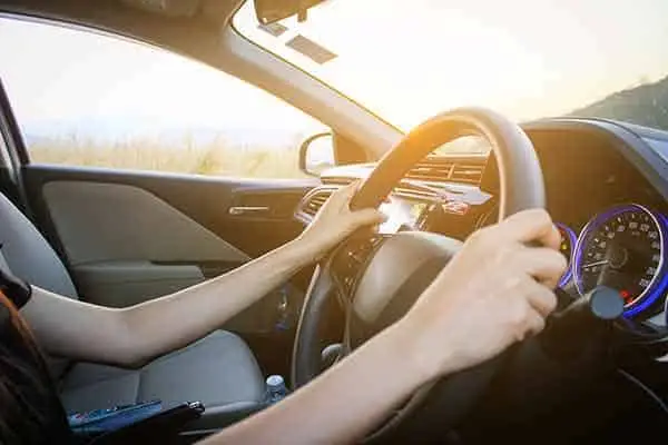 A driver's hands on the steering wheel with the low sun ahead on the road
