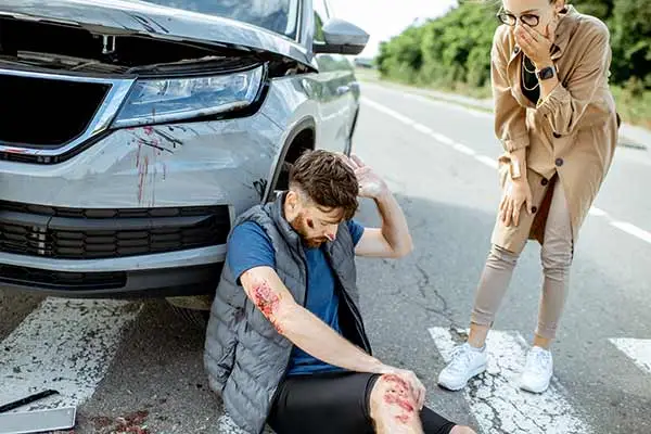 An injured man with bleeding arms sitting on the road by a car as a woman looks on in shock