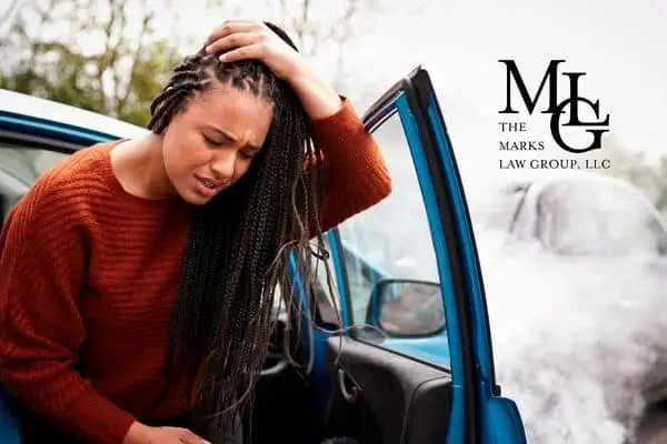 Woman holding her head in pain beside the open door of a damaged blue car