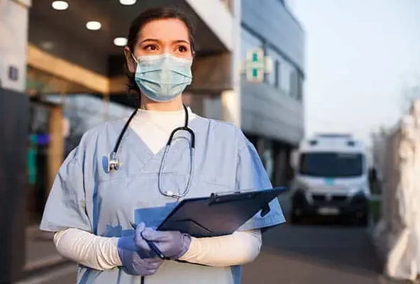 Masked nurse in scrubs holding a clipboard outside a hospital
