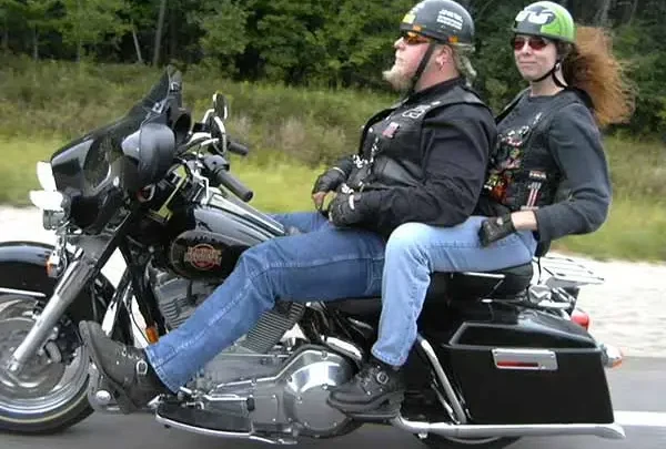 Two helmeted riders on a touring motorcycle along a wooded road