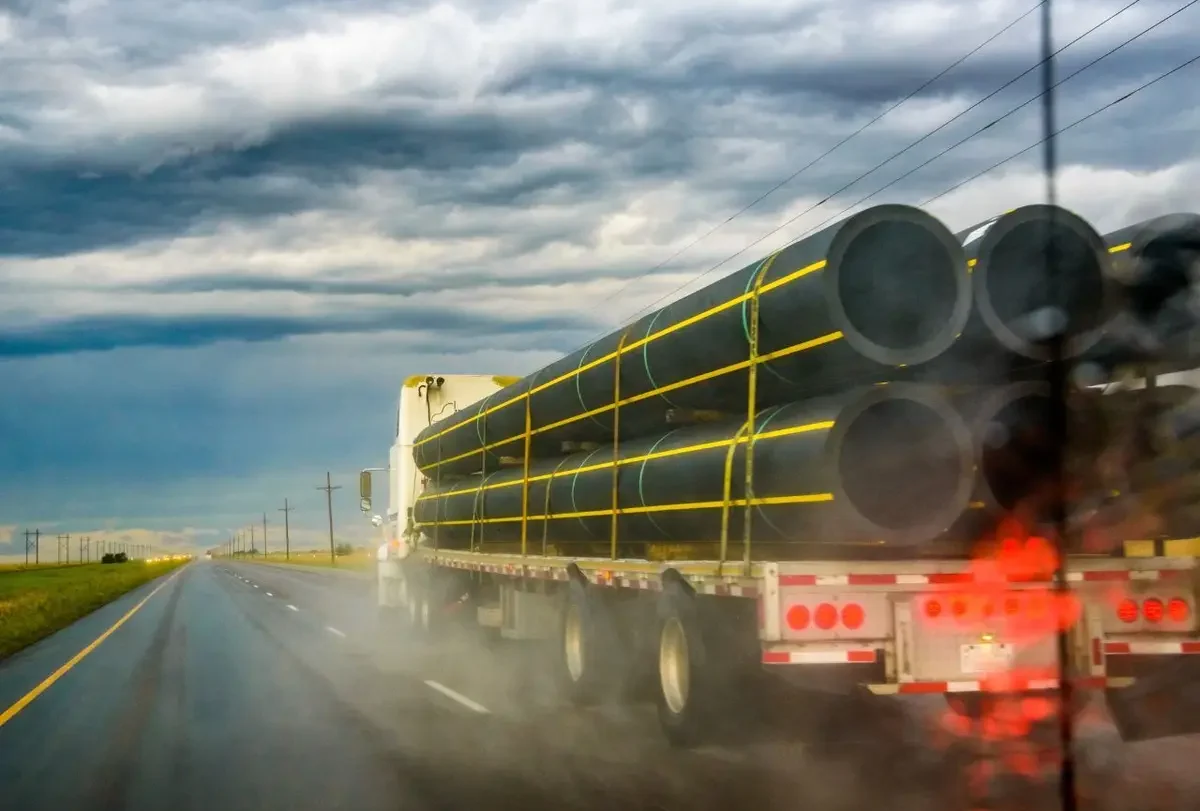 Semi truck hauling large pipes on a wet highway under storm clouds