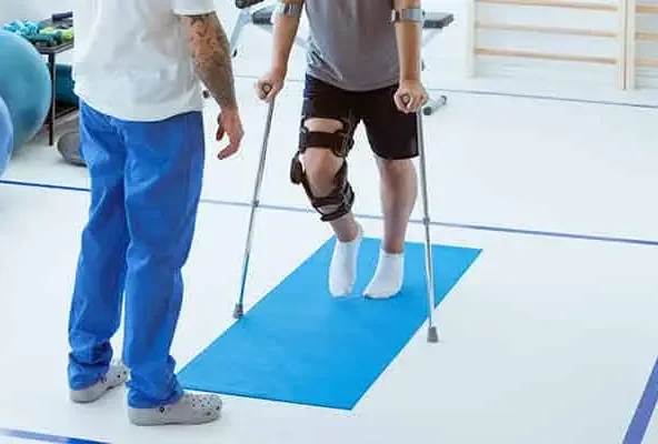 Patient with a leg brace walking on crutches beside a therapist