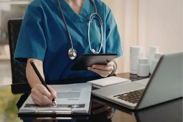 Nurse in scrubs writing on a clipboard while holding a tablet