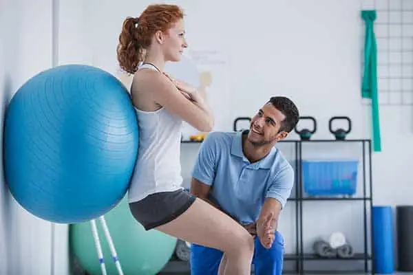 A physical therapist guiding a woman balancing against a large blue exercise ball