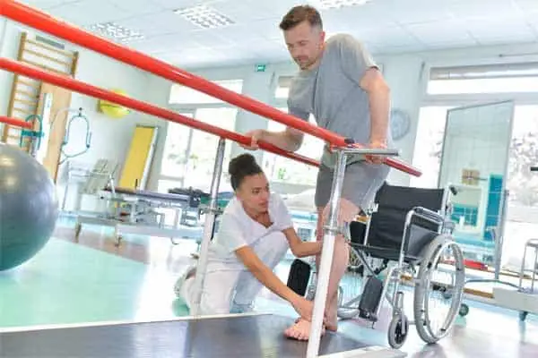 Physiotherapist steadying a man walking between parallel bars, wheelchair behind him