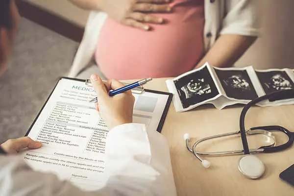 A doctor filling in a medical form beside a pregnant patient, ultrasound scans on the desk