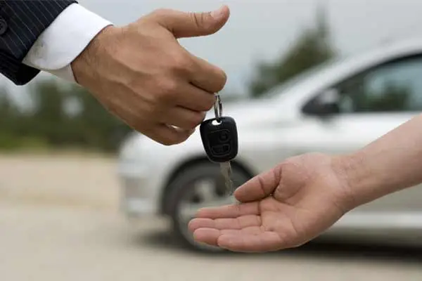 A hand in a suit passing a car key to an open palm in front of a white car