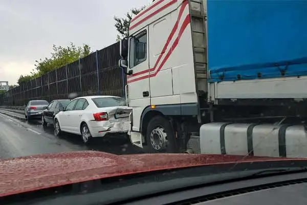 A white car rear-ended by a semi truck in slow traffic on a wet road