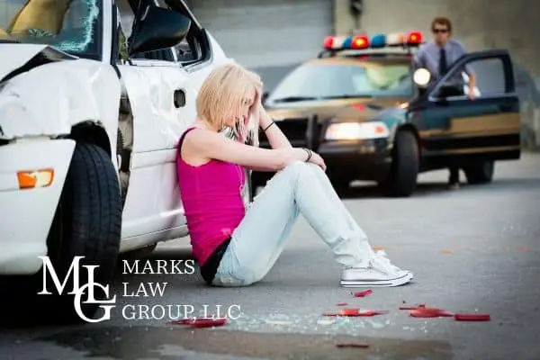 Young woman sitting on the ground against a wrecked car as a police officer approaches