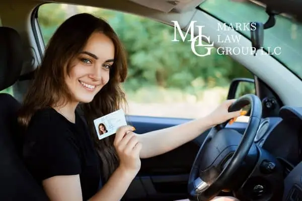 A young driver smiling in the driver's seat while holding up her license