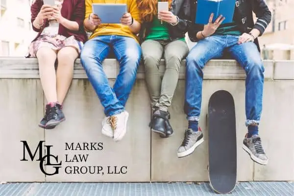 Four teenagers sitting on a wall with phones, a tablet and a book, a skateboard beside them