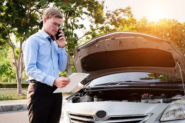 A man on the phone with a clipboard beside a car with its hood raised