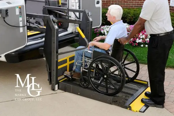 Older man in a wheelchair rolled onto a van's yellow lift ramp by an attendant