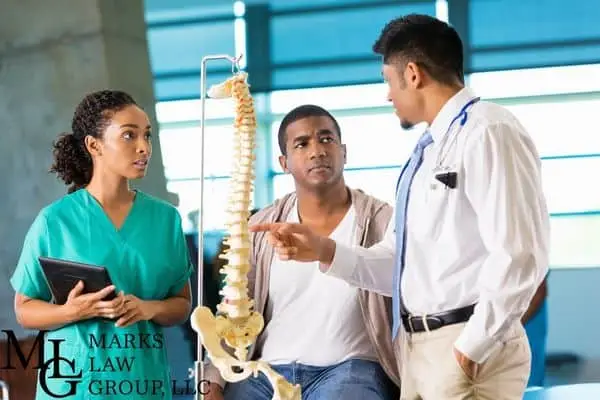 Two clinicians talking with a seated man beside a model of the human spine