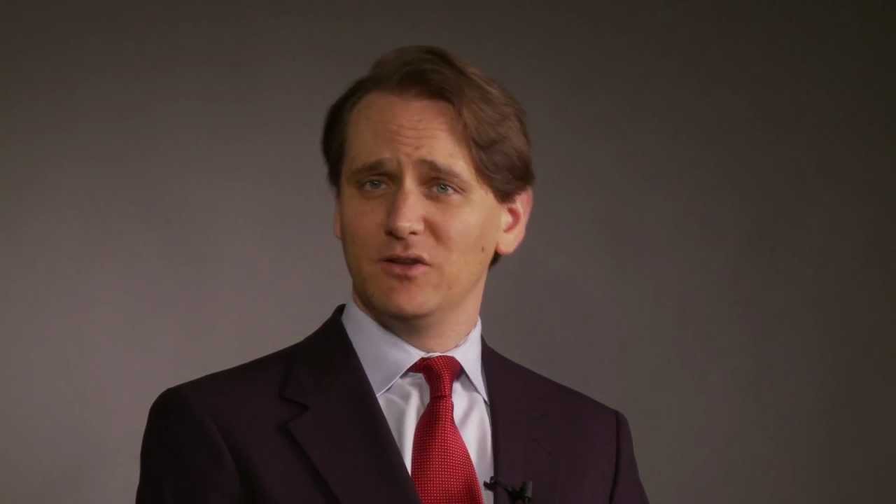 Man in a dark suit and red tie speaking against a plain grey backdrop