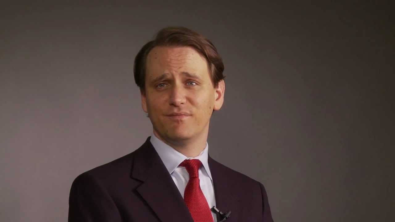Man in a dark suit and red tie looking toward the camera, grey backdrop